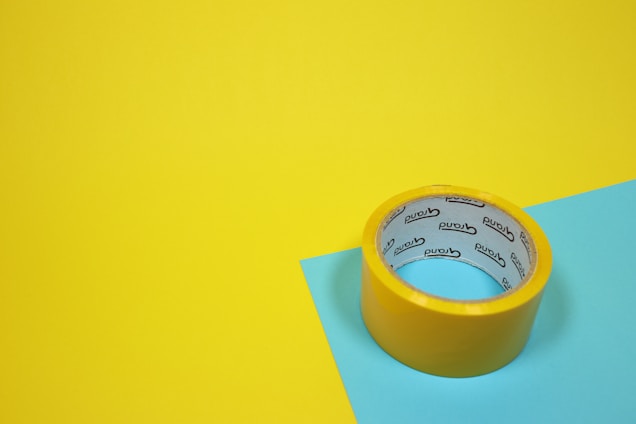 A roll of yellow adhesive tape sits on a yellow and blue surface. The tape has text on the inside.