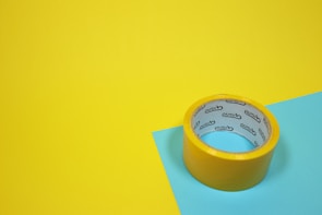A roll of yellow adhesive tape sits on a yellow and blue surface. The tape has text on the inside.