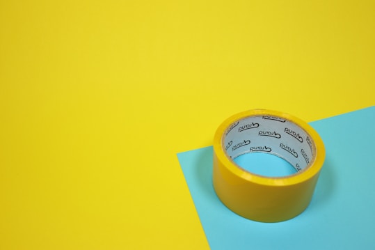 A roll of yellow adhesive tape sits on a yellow and blue surface. The tape has text on the inside.