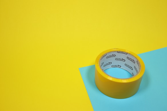 A roll of yellow adhesive tape sits on a yellow and blue surface. The tape has text on the inside.