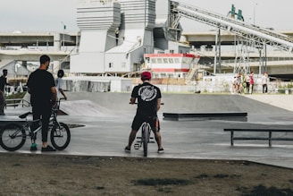 Several people are gathered at a skate park, with two individuals standing beside bikes and others in the background near ramps and benches. Industrial buildings and overpasses are visible, adding an urban backdrop to the scene.