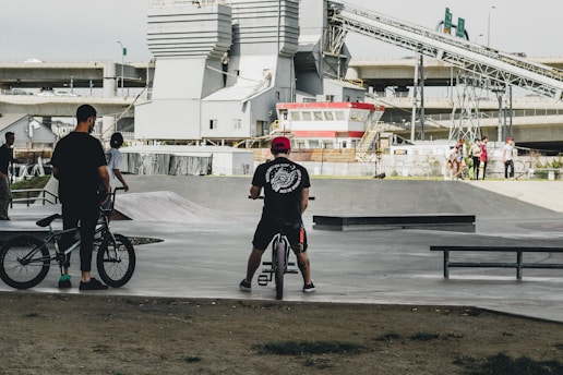 Several people are gathered at a skate park, with two individuals standing beside bikes and others in the background near ramps and benches. Industrial buildings and overpasses are visible, adding an urban backdrop to the scene.