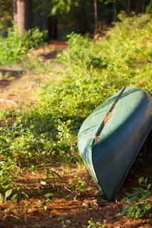 A green canoe is overturned and resting on a patch of grass surrounded by greenery and trees. Sunlight filters through the leaves, casting dappled shadows on the ground. The canoe appears to be unused and possibly stored in a natural setting, adding a peaceful and serene atmosphere.