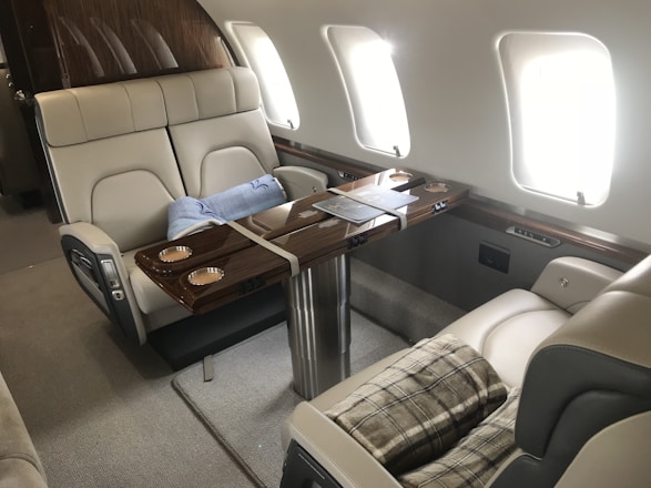 Luxurious interior of a private jet featuring two beige leather seats with a wooden table in between. The table has integrated cup holders and a small tray with reading material. A soft blue blanket is folded on one seat, and a plaid blanket is draped over the other.