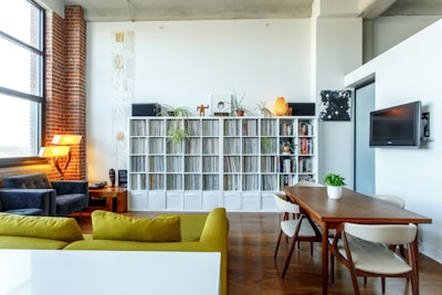 A modern living room features a bright and airy design with a large window allowing natural light to flood the space. The room includes a green sofa in the foreground, a wooden dining table with white chairs, and a cozy seating area with a dark armchair and a unique lamp. A tall shelf filled with vinyl records and plants adds a decorative touch, while a flat-screen TV is mounted on the wall.