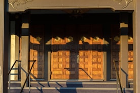 A set of ornate wooden doors with a geometric pattern, illuminated by warm sunlight. The doors are framed by a decorative architrave and flanked by a pair of handrails. Shadows cast intricate shapes across the surface of the doors.