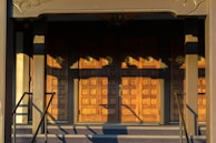 A set of ornate wooden doors with a geometric pattern, illuminated by warm sunlight. The doors are framed by a decorative architrave and flanked by a pair of handrails. Shadows cast intricate shapes across the surface of the doors.