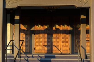 A set of ornate wooden doors with a geometric pattern, illuminated by warm sunlight. The doors are framed by a decorative architrave and flanked by a pair of handrails. Shadows cast intricate shapes across the surface of the doors.