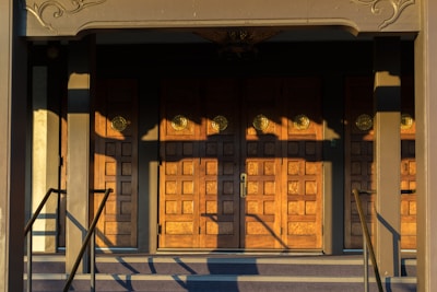 A set of ornate wooden doors with a geometric pattern, illuminated by warm sunlight. The doors are framed by a decorative architrave and flanked by a pair of handrails. Shadows cast intricate shapes across the surface of the doors.