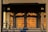 A set of ornate wooden doors with a geometric pattern, illuminated by warm sunlight. The doors are framed by a decorative architrave and flanked by a pair of handrails. Shadows cast intricate shapes across the surface of the doors.