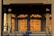 A set of ornate wooden doors with a geometric pattern, illuminated by warm sunlight. The doors are framed by a decorative architrave and flanked by a pair of handrails. Shadows cast intricate shapes across the surface of the doors.