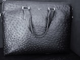 A black textured leather bag features a distinct pattern resembling an ostrich skin design. The bag has two handles and a brand name 'DA MILANO' printed on the upper center.