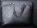 A black textured leather bag features a distinct pattern resembling an ostrich skin design. The bag has two handles and a brand name 'DA MILANO' printed on the upper center.