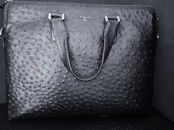 A black textured leather bag features a distinct pattern resembling an ostrich skin design. The bag has two handles and a brand name 'DA MILANO' printed on the upper center.