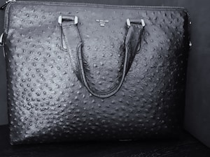 A black textured leather bag features a distinct pattern resembling an ostrich skin design. The bag has two handles and a brand name 'DA MILANO' printed on the upper center.