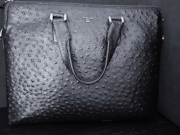 A black textured leather bag features a distinct pattern resembling an ostrich skin design. The bag has two handles and a brand name 'DA MILANO' printed on the upper center.