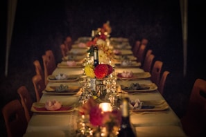 A long dining table is set for an evening event, adorned with elegant place settings, decorative floral centerpieces, and wine bottles. Soft, ambient lighting from candles creates a warm and inviting atmosphere.