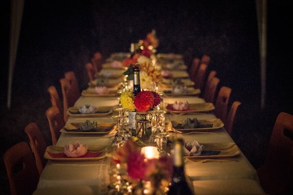 A long dining table is set for an evening event, adorned with elegant place settings, decorative floral centerpieces, and wine bottles. Soft, ambient lighting from candles creates a warm and inviting atmosphere.