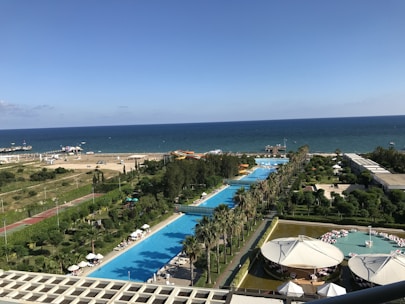 A coastal resort area featuring a long stretch of swimming pools lined with palm trees, adjacent to a clear blue sea. The foreground includes a poolside area with people relaxing under umbrellas, while further towards the horizon, there is a beach with a few parked cars and brightly colored inflatables. The scene is vibrant with lush greenery and well-maintained gardens surrounding the resort facilities.