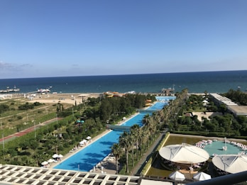 A coastal resort area featuring a long stretch of swimming pools lined with palm trees, adjacent to a clear blue sea. The foreground includes a poolside area with people relaxing under umbrellas, while further towards the horizon, there is a beach with a few parked cars and brightly colored inflatables. The scene is vibrant with lush greenery and well-maintained gardens surrounding the resort facilities.