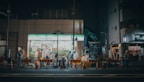 A nighttime construction scene in front of a FamilyMart convenience store. There are construction workers in high-visibility clothing, orange and white traffic cones, and barriers marking the work area. A small excavator is also visible, along with various construction equipment and tools. The store has bright fluorescent lighting, contrasting with the dim street.