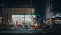 A nighttime construction scene in front of a FamilyMart convenience store. There are construction workers in high-visibility clothing, orange and white traffic cones, and barriers marking the work area. A small excavator is also visible, along with various construction equipment and tools. The store has bright fluorescent lighting, contrasting with the dim street.