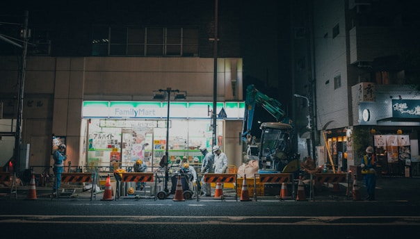 A nighttime construction scene in front of a FamilyMart convenience store. There are construction workers in high-visibility clothing, orange and white traffic cones, and barriers marking the work area. A small excavator is also visible, along with various construction equipment and tools. The store has bright fluorescent lighting, contrasting with the dim street.