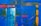An abstract painting featuring a vibrant blue background with dynamic strokes of orange and red. Vertical and horizontal lines intersect across the canvas, adding structure and depth. A splash of white creates a focal point, contrasted by the bold colors around it.