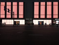 A large store facade illuminated with pink light panels, showcasing the name 'Victoria's Secret' prominently in the center. The windows display mannequins and advertisements. Several people are walking along the street in silhouette.