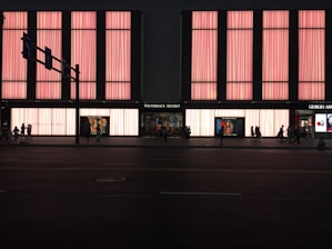 A large store facade illuminated with pink light panels, showcasing the name 'Victoria's Secret' prominently in the center. The windows display mannequins and advertisements. Several people are walking along the street in silhouette.