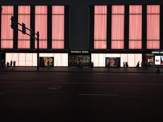A large store facade illuminated with pink light panels, showcasing the name 'Victoria's Secret' prominently in the center. The windows display mannequins and advertisements. Several people are walking along the street in silhouette.