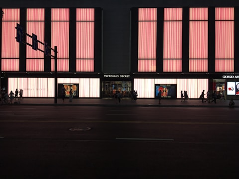 A large store facade illuminated with pink light panels, showcasing the name 'Victoria's Secret' prominently in the center. The windows display mannequins and advertisements. Several people are walking along the street in silhouette.