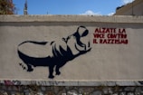 A street art piece on a concrete wall featuring a silhouette of a large animal, possibly a hippo, with a person inside shaped as if they are raising their arm. There is text in bold red letters that reads 'Alzate la Voce Contro il Razzismo,' which translates to 'Raise your voice against racism.' Small graffiti tags are visible at the bottom of the wall.