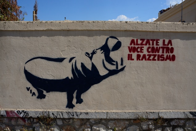 A street art piece on a concrete wall featuring a silhouette of a large animal, possibly a hippo, with a person inside shaped as if they are raising their arm. There is text in bold red letters that reads 'Alzate la Voce Contro il Razzismo,' which translates to 'Raise your voice against racism.' Small graffiti tags are visible at the bottom of the wall.