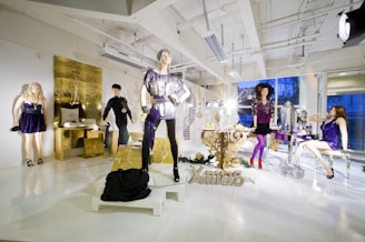 A brightly lit room with mannequins wearing fashionable clothing in a festive display. One mannequin wears a shiny silver top and black pants, while another dons a purple dress. A gold thematic decor surrounds them, including a table, candles, and a large glittery 'Xmas' sign. The windows in the back reveal a blue-tinted evening sky, and various decorative elements such as gift boxes, mirrors, and ornaments are present.