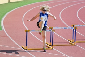 An athlete with blonde hair is mid-air attempting to clear a hurdle on a running track. Wearing a blue and white athletic outfit with the number 135, the runner has sunglasses on and appears focused and determined. The bright red track contrasts with the hurdles that have blue frames and yellow bases.