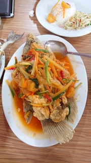 A plate of fried fish topped with a colorful sweet and sour sauce containing sliced vegetables and pineapple. Next to it, there is a plate with rice shaped into a dome, accompanied by a slice of pineapple and a side salad. The dishes are served on a wooden tabletop.