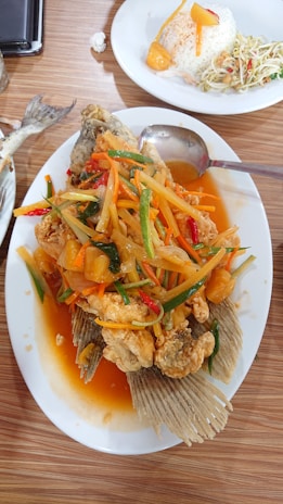 A plate of fried fish topped with a colorful sweet and sour sauce containing sliced vegetables and pineapple. Next to it, there is a plate with rice shaped into a dome, accompanied by a slice of pineapple and a side salad. The dishes are served on a wooden tabletop.