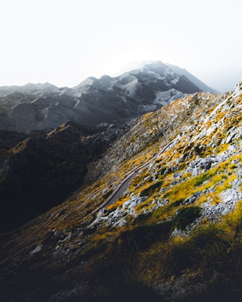 A rugged mountain landscape with steep rocky slopes. A narrow hiking trail winds its way through the terrain, surrounded by patches of greenery and yellowish vegetation. The distant mountains are shrouded in mist, adding a sense of mystery and depth to the scene.