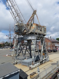 A large, weathered industrial crane with rust patches stands on a dock. In the background, there is a historical sailing ship with tall masts. The setting appears to be a maritime area with buildings and parked cars visible in the distance under a partly cloudy sky.