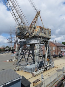 A large, weathered industrial crane with rust patches stands on a dock. In the background, there is a historical sailing ship with tall masts. The setting appears to be a maritime area with buildings and parked cars visible in the distance under a partly cloudy sky.