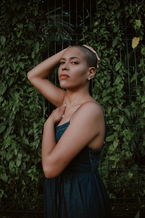 A person with a confident expression stands against a backdrop of lush green foliage. The individual is wearing a sleeveless dark blue dress and has a shaved head. One hand rests on the head while the other touches the chest, creating a composed and introspective pose.