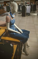 A mannequin dressed in a denim dress with a striped shirt underneath is seated on a wooden bench in a clothing store. Around the mannequin are neatly arranged clothes, and various racks filled with garments are visible in the background under bright lighting.