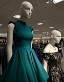 Mannequins dressed in elegant gowns on display in a retail store. One mannequin is wearing a sleeveless teal dress with ruffled shoulders, while the other is in a black lace off-shoulder gown. The store has a well-lit ceiling with numerous lights and carries a variety of clothing items visible in the background.