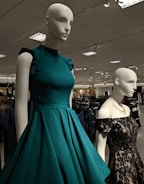 Mannequins dressed in elegant gowns on display in a retail store. One mannequin is wearing a sleeveless teal dress with ruffled shoulders, while the other is in a black lace off-shoulder gown. The store has a well-lit ceiling with numerous lights and carries a variety of clothing items visible in the background.