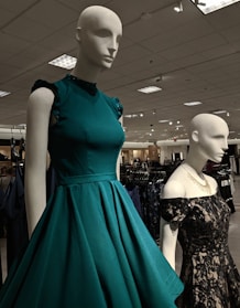 Mannequins dressed in elegant gowns on display in a retail store. One mannequin is wearing a sleeveless teal dress with ruffled shoulders, while the other is in a black lace off-shoulder gown. The store has a well-lit ceiling with numerous lights and carries a variety of clothing items visible in the background.