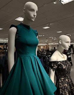 Mannequins dressed in elegant gowns on display in a retail store. One mannequin is wearing a sleeveless teal dress with ruffled shoulders, while the other is in a black lace off-shoulder gown. The store has a well-lit ceiling with numerous lights and carries a variety of clothing items visible in the background.