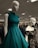 Mannequins dressed in elegant gowns on display in a retail store. One mannequin is wearing a sleeveless teal dress with ruffled shoulders, while the other is in a black lace off-shoulder gown. The store has a well-lit ceiling with numerous lights and carries a variety of clothing items visible in the background.