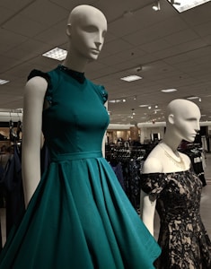 Mannequins dressed in elegant gowns on display in a retail store. One mannequin is wearing a sleeveless teal dress with ruffled shoulders, while the other is in a black lace off-shoulder gown. The store has a well-lit ceiling with numerous lights and carries a variety of clothing items visible in the background.