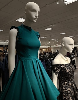 Mannequins dressed in elegant gowns on display in a retail store. One mannequin is wearing a sleeveless teal dress with ruffled shoulders, while the other is in a black lace off-shoulder gown. The store has a well-lit ceiling with numerous lights and carries a variety of clothing items visible in the background.